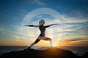 Silhouette of young woman practicing yoga standing in warrior pose on the beach