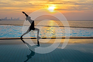 Silhouette young woman practicing yoga on swimming pool and the beach at sunset