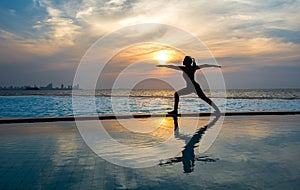 Silhouette young woman practicing yoga on swimming pool and the beach at sunset.