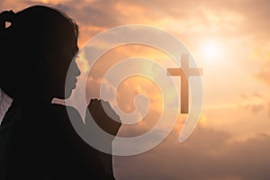 Silhouette of young woman praying with a cross at sunrise, Christian Religion concept background