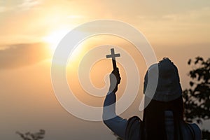 Silhouette of young woman praying with a cross at sunrise, Christian Religion concept background