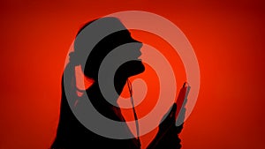 Silhouette of young woman put on ear-phones and listen to music on smart phone on red background