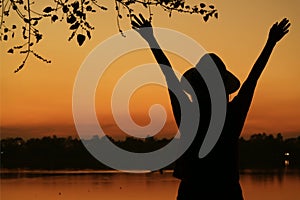 Silhouette of a young woman raising arms against beautiful orange color sunset sky on the lake shore