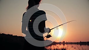 Silhouette of a young woman resting on nature fishing on a fishing rod by spinning at dawn