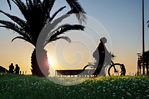 Silhouette of a young woman riding the bike at seaside park at sunset with field foreground