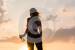 Silhouette of young woman in the sport clothes with bottle of clear mineral water on the sunset sky backgrounds. The concept