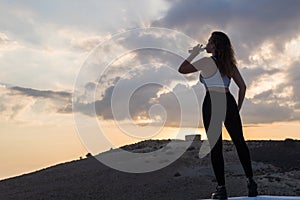 Silhouette of young woman in the sport clothes drinking mineral water from bottle on the sunset sky and mountain backgrounds.