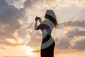 Silhouette young woman in the sport clothes, headphones and mobile phone  on the sunset sky backgrounds. The concept of healthy
