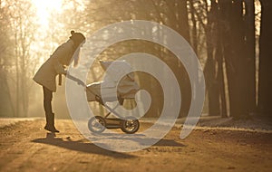 Silhouette of young woman on sunny footpath and looking at child in baby stroller. Sunny winter park, childcare and love