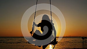 Silhouette of young woman swinging on a swing through the sun at beautiful sunset on tropical beach. slow motion