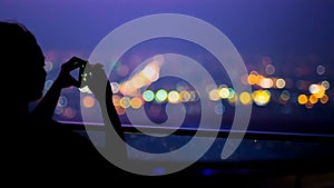 Silhouette young woman taking photo with smartphone to beautiful colors bokeh of city night light from top of building at bangkok