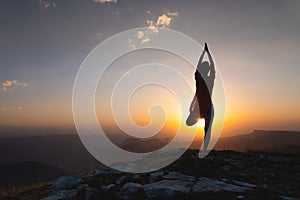 Silhouette of a young woman in a tree pose outdoors in the mountains watching the sunset, yoga balance near the cliff