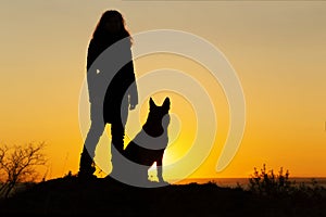 Silhouette of young woman walking with a dog in the field at sunset, pet sitting near girl`s leg on nature, figure of friends in