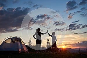 Silhouette of young woman and child giving each other high five near camping at dawn on top of mountain