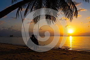 Silhouetted couple in love walks on the beach during sunset. Riding on a swing tied to a palm tree and watching the sun go down