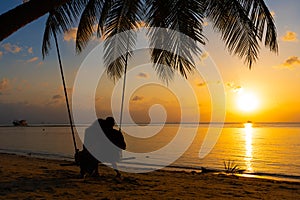 Silhouetted couple in love walks on the beach during sunset. Riding on a swing tied to a palm tree and watching the sun go down