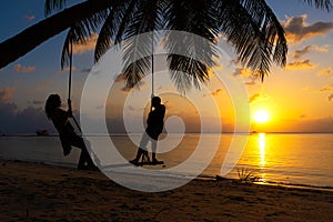 Silhouetted couple in love walks on the beach during sunset. Riding on a swing tied to a palm tree and watching the sun go down