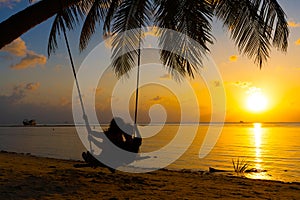 Silhouetted couple in love walks on the beach during sunset. Riding on a swing tied to a palm tree and watching the sun go down