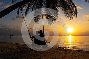 Silhouetted couple in love walks on the beach during sunset. Riding on a swing tied to a palm tree and watching the sun go down