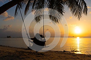 Silhouetted couple in love walks on the beach during sunset. Riding on a swing tied to a palm tree and watching the sun go down