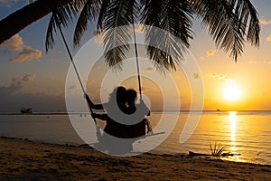 Silhouetted couple in love walks on the beach during sunset. Riding on a swing tied to a palm tree and watching the sun go down