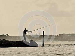 Silhouetted fisherman casting a line from a rocky pier at hazy sunset.