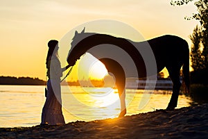 Silhouetted girl walking at sunset with a horse by the lake