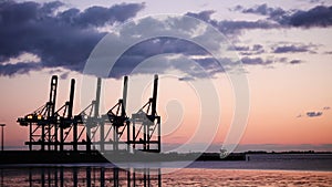 Silhouetted tanker cargo ship boat sailing into a port with cranes at sunset or sunrise