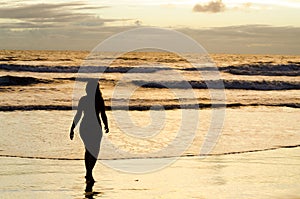 A silhouetted woman walking near the sea on a beach with the sun rising and the sunrays reflecting in the sea water.