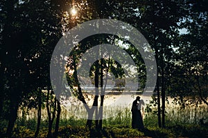 Silhouettes of bride and groom are kissing on the shore of lake between trees at sunset