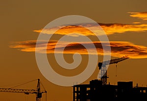 Silhouettes of construction cranes and construction on sunset sky background. Yellow hot clouds in the sky above