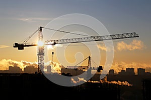 Silhouettes of construction cranes and unfinished residential buildings against the sky and shining sun