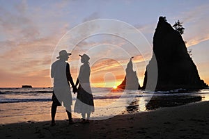 Silhouettes of couple holding hands on beach by sea stack rocks at sunset.