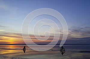 Silhouettes of cyclists at sunset of the day on the sandy bank of the river. Riding on bicycles on the water