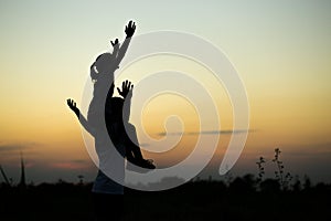 Silhouettes of father and daughter on his shoulders with hands up having fun, against sunset sky.