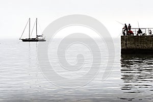 Silhouettes of fishermen on the pier on the background of the silhouette of a yacht on the calm sea.