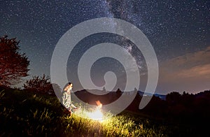 Silhouettes of girl near haystack and bonfire under starry sky on which Milky Way is visible