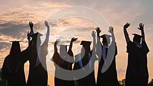 Silhouettes of graduates in black robes wave their arms against the evening sunset and toss their caps.