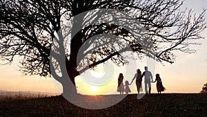 Silhouettes of happy family walking in the meadow near a big tree during sunset.