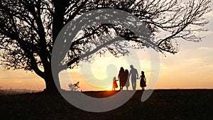 Silhouettes of happy family walking in the meadow near a big tree during sunset.