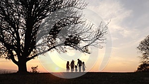 Silhouettes of happy family walking in the meadow near a big tree during sunset.