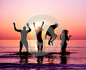 Silhouettes of happy friends jumping inside water on the beach at sunset - Group of young people having fun on summer vacation -