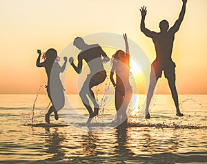 Silhouettes of happy friends jumping inside water on the beach at sunset - Group of young people having fun on summer vacation -