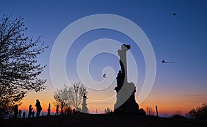 Silhouettes of kids flying kites with stone christianity monument in the foreground at sunset