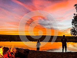 Silhouettes of a man and a child fishing on the coast of a lake at sunset with a bonfire behind them