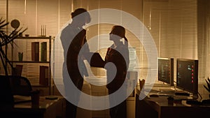 Silhouettes of a man and a woman shake hands and discuss the report. The managers works in the evening in a dark office