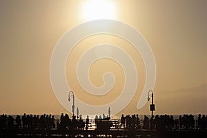 Silhouettes of many unrecognizable people walking across the Santa Monica Pier at a slightly foggy sunset