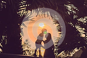 Silhouettes of mother and son, who meet the sunset in the tropics against the backdrop of palm trees