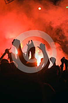 Silhouettes of people against the red light from the torch in front of the stage at a music rock festival. A crowd of fans at a