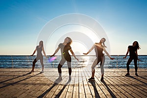 Silhouettes of sportive girls dancing zumba near sea at sunrise.